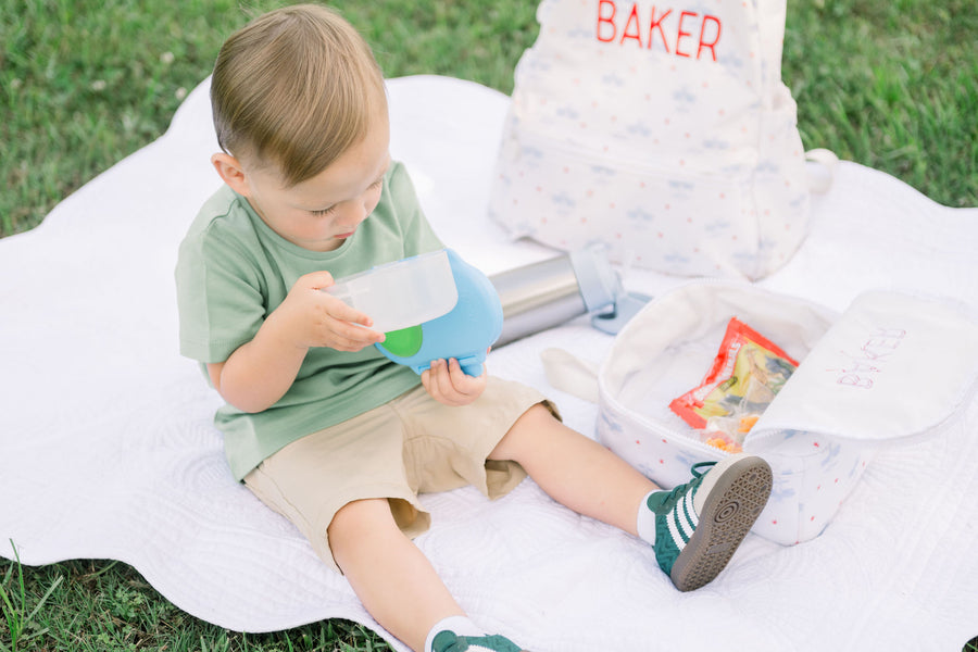 Toddler boy sitting on a blanket next to a backpack, lunch bag and water bottle opening the snack box.