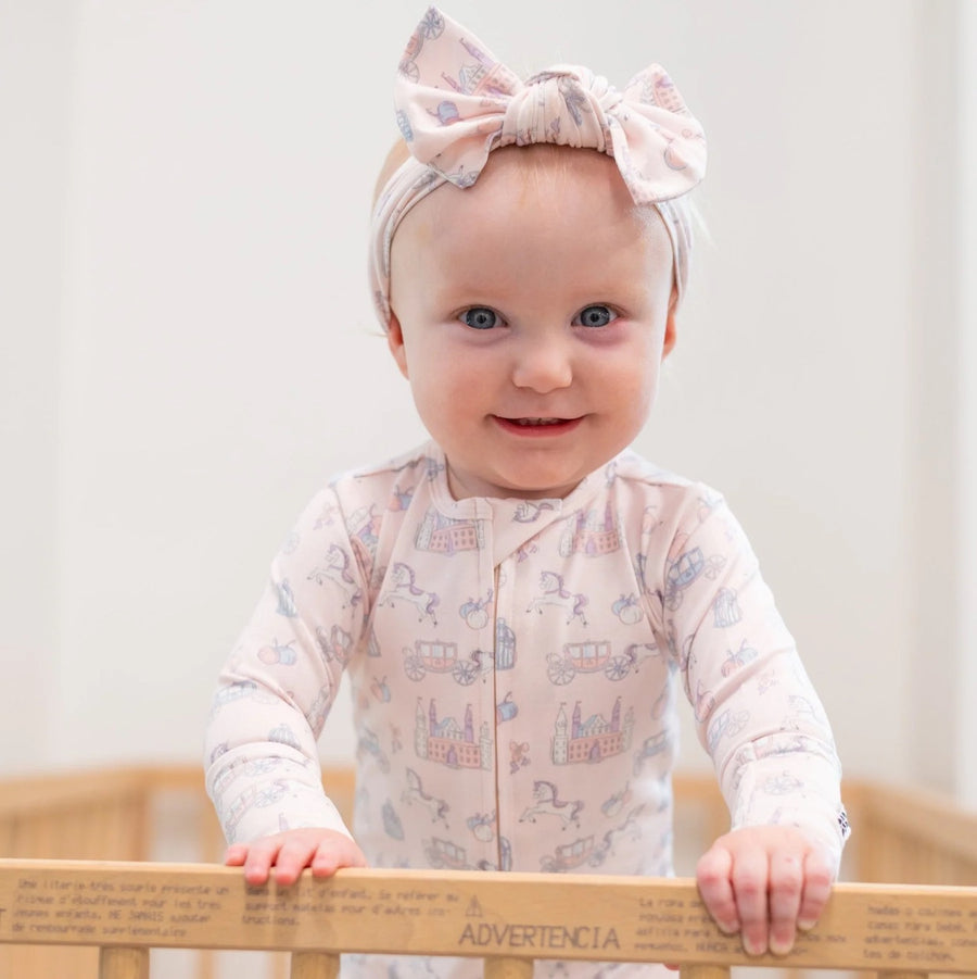 Baby girl standing up in a crib with a bow on her head, wearing the bamboo zipper romper in the Fairy Godmother Print.